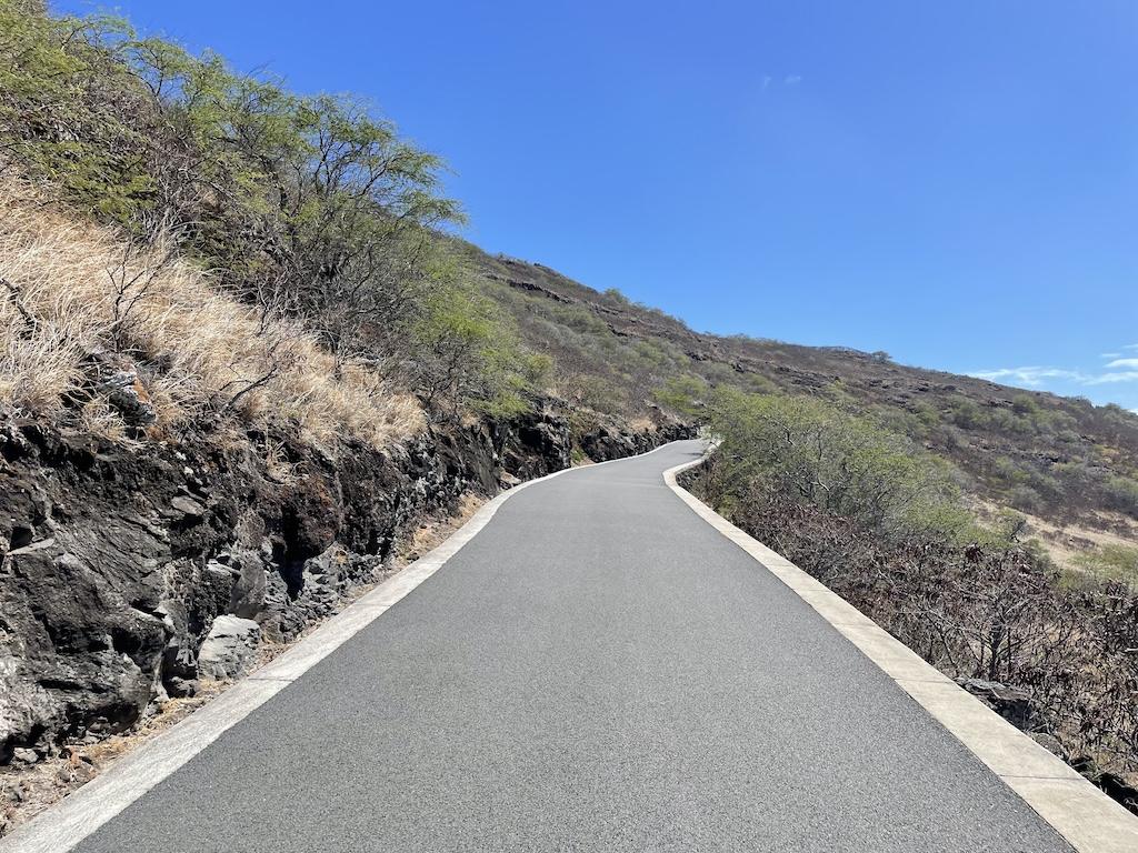 The Makapu'u Lighthouse Hike with smooth paved trail on an incline and volcanic rock and shrubs on the side of the trail