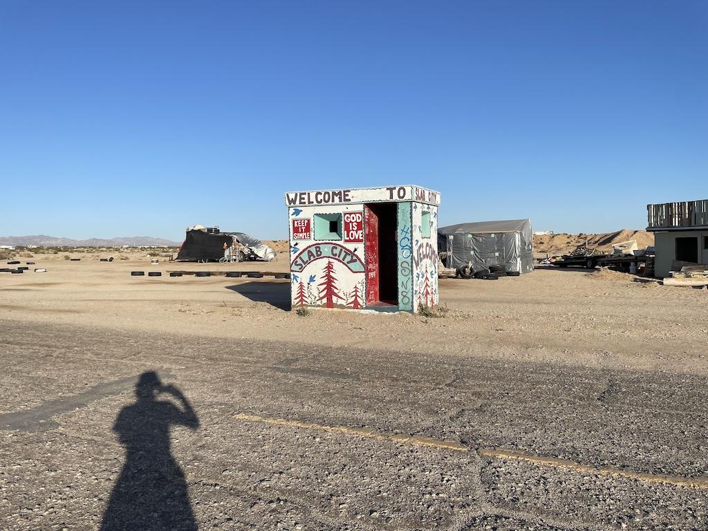 Entrance booth Luna saw while visiting Slab City with Luna’s shadow in front and buildings in the distance in Niland, California
