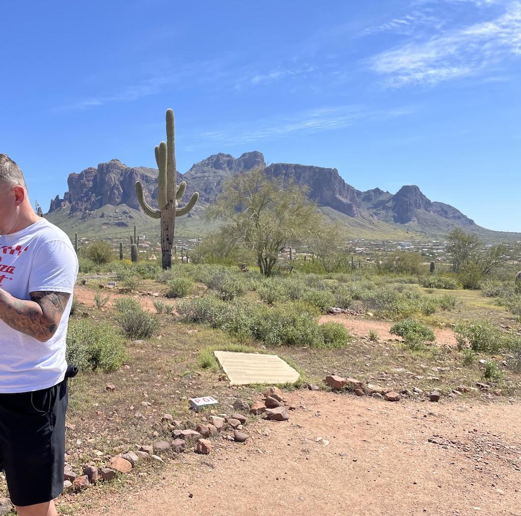 View of Superstition Mountains in background with man on the side taken from the Lost Dutchman museum in Arizona