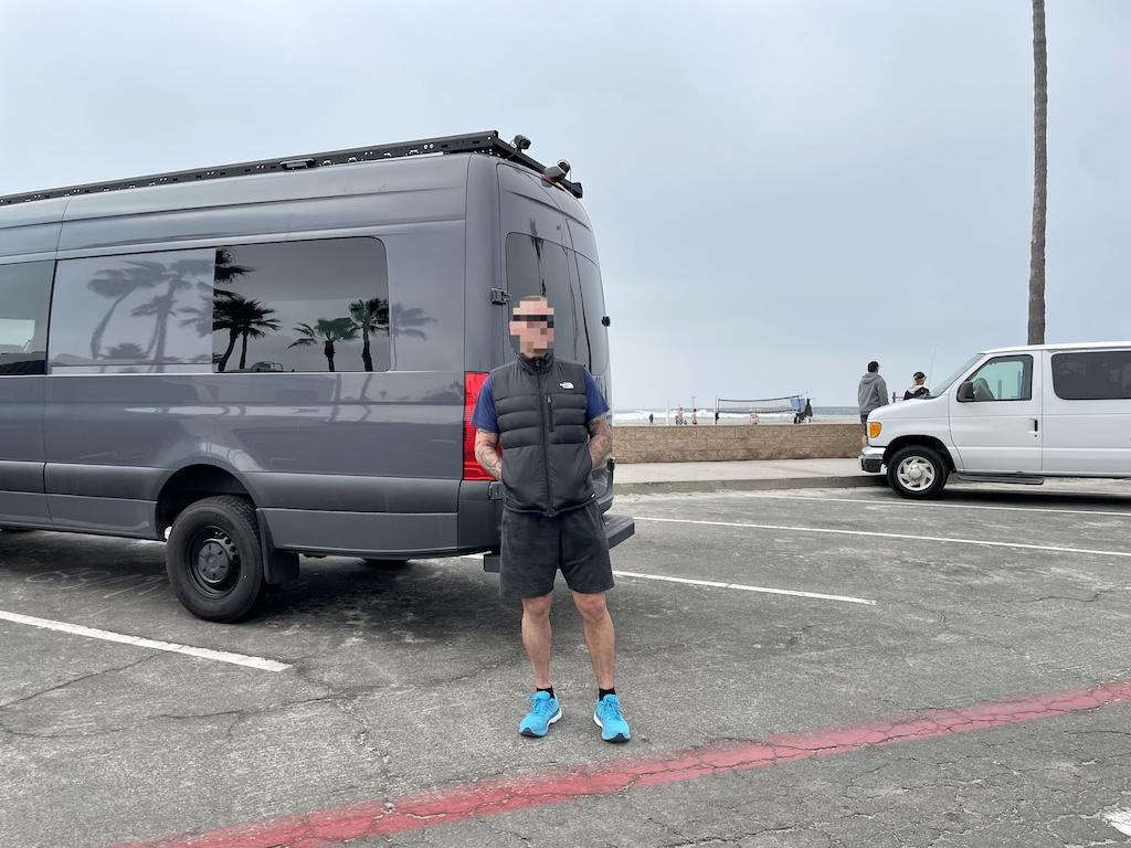 View of man standing outside Luna’s Sprinter camper van in the overnight parking lot in Oceanside Harbor, California