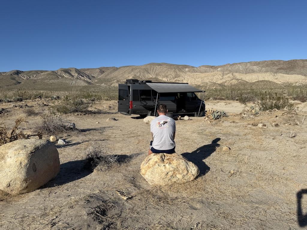 Man sitting on rock, looking at Sprinter camper van off Coyote Canyon Road, at Anza-Borrego Desert State Park, California.