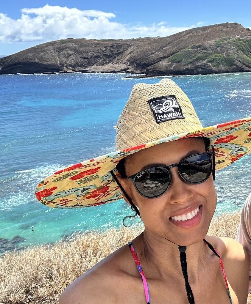 Close up of Luna smiling to the camera wearing a straw hat with the ocean in the background at Hanauma Bay in Oahu, Hawaii.